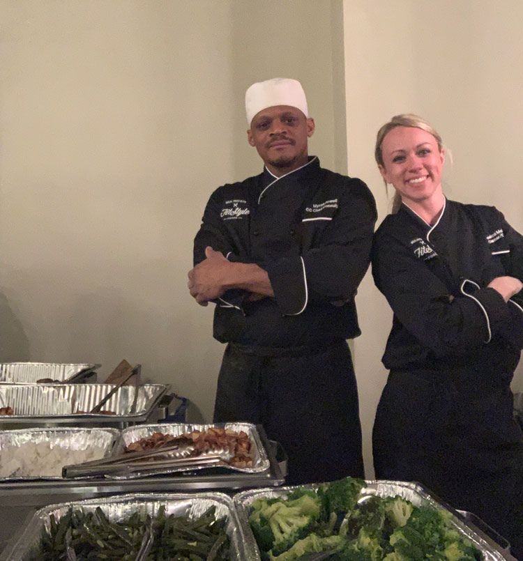 Jessica McLead, owner of Fitstyle Foods, and Chef Myron standing side by side behind buffet table covered in catering trays